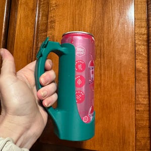 Puede incluir: Un soporte para latas de color verde azulado con un asa, que sostiene una lata rosa y roja. La lata tiene texto que dice "200mg Caffeine" y otros gr&aacute;ficos. El soporte est&aacute; dise&ntilde;ado para encajar perfectamente en la lata, proporcionando un agarre c&oacute;modo.