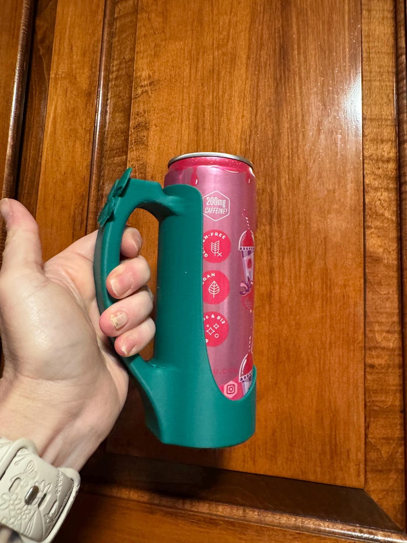 Puede incluir: Un soporte para latas de color verde azulado con un asa, que sostiene una lata rosa y roja. La lata tiene texto que dice "200mg Caffeine" y otros gr&aacute;ficos. El soporte est&aacute; dise&ntilde;ado para encajar perfectamente en la lata, proporcionando un agarre c&oacute;modo.