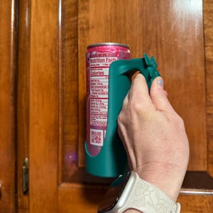 Puede incluir: Un soporte para latas de color verde azulado con un asa, dise&ntilde;ado para sujetar una lata de bebida. La lata es rosa y blanca. El soporte lo sostiene una persona. El fondo es un armario de madera.