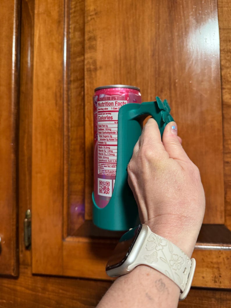 Puede incluir: Un soporte para latas de color verde azulado con un asa, dise&ntilde;ado para sujetar una lata de bebida. La lata es rosa y blanca. El soporte lo sostiene una persona. El fondo es un armario de madera.