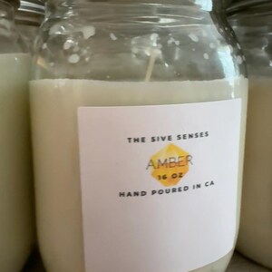 May include: A clear glass jar candle with a cream-coloured wax. The label reads "THE SIVE SENSES AMBER 16 OZ HAND POURED IN CA". The candle is in a clear glass jar with a metal lid. The candle is part of a set.