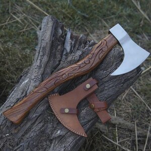 May include: A handcrafted axe with a wooden handle featuring carved details. The axe head is silver, and a brown leather sheath is included. The axe is displayed on a weathered wooden surface, showcasing its rustic design.