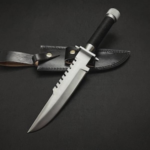May include: A survival knife with a silver blade and a black handle, resting in a black leather sheath. The knife has a serrated edge and a silver metal guard. The handle is wrapped in black material, and the sheath has a belt loop.