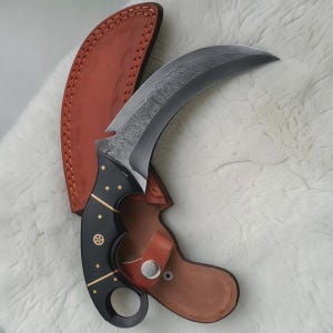 May include: A curved karambit knife with a patterned blade and a black handle adorned with gold accents. The knife is paired with a brown leather sheath. The blade has a unique, textured pattern.