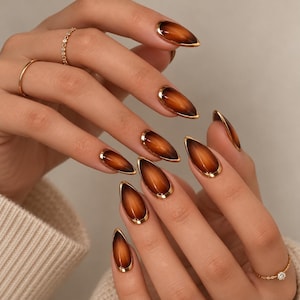 May include: Almond-shaped press-on nails featuring a deep brown gradient finish with a reflective cat-eye effect and a metallic gold border. The set includes two thin gold rings with small clear stones worn on the fingers.