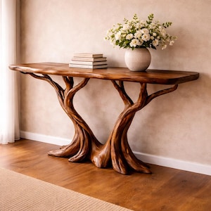 Op de afbeelding: Een handgemaakte houten consoletafel met een natuurlijke boomstamvoet en een blad met levende rand. De tafel is warmbruin gebeitst en is versierd met een vaas met witte bloemen en een stapel boeken. De tafel is ongeveer 183 cm lang.