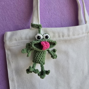 May include: A handmade green crochet frog with a pink tongue and white googly eyes is attached to a white canvas tote bag. The frog has small arms and legs. The background is a solid purple color.