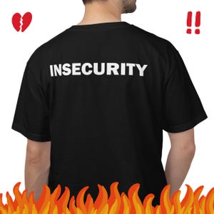 以下が含まれることがあります： 背面に白い大文字でINSECURITYという単語がプリントされた黒い半袖Tシャツ。画像には、左上に赤い壊れたハートのアイコン、右側に2つの赤い感嘆符、そして下部にオレンジ色の炎が描かれています。