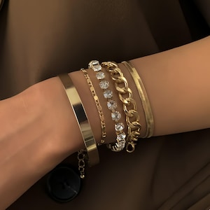 Op de afbeelding: Een verzameling gouden armbanden in verschillende stijlen. De armbanden omvatten een effen armband, een fijne ketting, een streng met heldere kristallen, een dikke ketting en een getextureerde band. De armbanden zijn gestapeld op een pols tegen een bruine achtergrond.