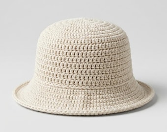 May include: A solid cream-colored bucket hat crafted with a dense, uniform crochet stitch pattern. The hat features a rounded crown and a short, downward-sloping brim that encircles the entire base, maintaining a consistent texture throughout.