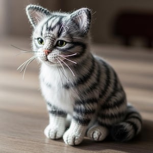 Può includere: Un gattino all'uncinetto con strisce grigie e bianche, occhi verdi e lunghi baffi bianchi. Il gatto fatto a mano è seduto su una superficie di legno. Le orecchie del gatto sono a punta e le zampe sono bianche.