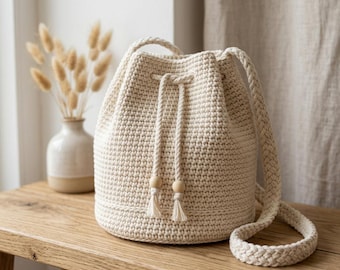May include: A cream-colored crochet bucket bag featuring a thick braided shoulder strap and a drawstring closure with two dangling cords finished with small wooden beads. The bag has a structured cylindrical base and a dense, uniform stitch pattern.