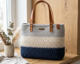 May include: A rectangular crochet tote bag featuring horizontal color-blocked stripes in light gray, cream, and navy blue. The bag has two tan leather handles attached with brass-colored metal rivets and a small rectangular tag labeled HANDMADE.