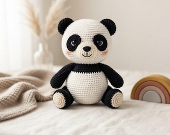 Puede incluir: Un peluche de panda tejido a ganchillo con un cuerpo redondo blanco, extremidades negras, orejas negras y manchas negras alrededor de los ojos. El panda tiene una pequeña nariz negra, una boca bordada y mejillas rosadas, hecho con hilo.