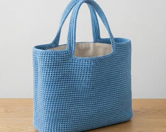 May include: A light blue crochet tote bag featuring a structured rectangular body and two matching top handles. The exterior is crafted with a dense, uniform stitch pattern, while the interior is lined with a plain, light-colored fabric.