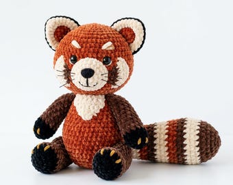 Puede incluir: Un juguete de peluche de panda rojo hecho a ganchillo, con una cabeza redonda con detalles en color crema, orejas de color marrón oscuro y patas negras. El cuerpo es de un cálido naranja óxido y la cola larga y gruesa muestra rayas alternas en crema, óxido y marrón oscuro.