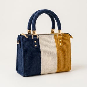 May include: A rectangular crochet handbag featuring vertical color-blocked stripes in navy blue, cream, and mustard yellow. The bag includes two rounded navy blue crochet handles attached with gold-toned metal hardware and a top zipper closure.