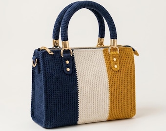 May include: A rectangular crochet handbag featuring vertical color-blocked stripes in navy blue, cream, and mustard yellow. The bag includes two rounded navy blue crochet handles attached with gold-toned metal hardware and a top zipper closure.