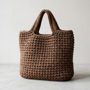 May include: A medium brown tote bag crafted with a thick, textured crochet knit pattern. The bag features a rectangular body with a flat base and two integrated, sturdy handles at the top, showcasing a consistent, dense stitch throughout the design.