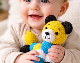 May include: A smiling baby holds a colorful, handmade crochet toy. The toy is a bear-like creature with a yellow head, black ears, and a blue and white body. The toy has a smiling face with a black nose and eyes. The baby is wearing a light-colored onesie.