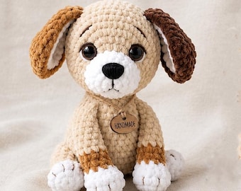 Puede incluir: Un perro de peluche tejido a ganchillo con cuerpo color canela, hocico y patas blancas. Tiene grandes ojos negros, nariz negra y orejas caídas en marrón claro y oscuro. Una pequeña etiqueta de madera en el cuello lleva la palabra HANDMADE.