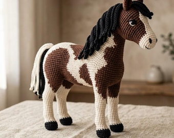 Puede incluir: Un juguete de caballo tejido a ganchillo con un cuerpo de patrón marrón y blanco, crin negra y cola negra. El caballo tiene una mancha blanca en la cara, ojos oscuros y cascos negros. La estructura del hilo es visible en toda la figura.