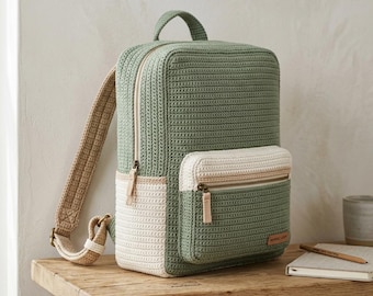 Puede incluir: Una mochila de ganchillo en color verde salvia y crema que presenta un compartimento principal rectangular, un bolsillo frontal más pequeño con cremallera, un asa superior y correas de hombro tejidas en color beige con herrajes metálicos.