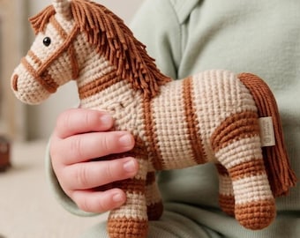 May include: A crocheted toy horse in tan and brown stripes. The horse has a brown mane and tail, and a tan bridle. The toy is held in a child's hands. The toy is a soft, handmade item.