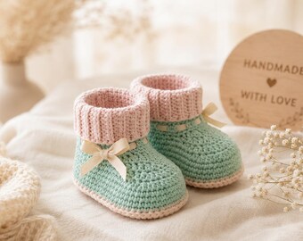 May include: A pair of crochet baby booties featuring a mint green lower section and a ribbed light pink cuff. Each bootie is accented with a small cream-colored ribbon bow tied at the front. The booties are crafted with a visible knit texture.