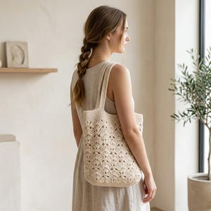 Pode incluir: Uma bolsa tote de crochê na cor creme com um padrão de pontos repetidos em forma de leque. A bolsa possui duas alças de ombro integradas e um corpo retangular feito de fio, exibindo um design texturizado e vazado em todo o exterior.