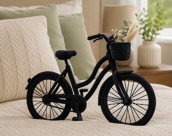 May include: A black crocheted bicycle figurine featuring a front basket filled with small dried flowers. The bicycle has detailed wheels with thin spokes, a seat, handlebars, and pedals, all crafted from black yarn in a dense, textured stitch.