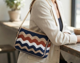 Puede incluir: Un bolso de mano rectangular de ganchillo con un patrón ondulado en azul marino, blanco y naranja terracota. El bolso cuenta con una correa de cadena de metal dorado y un cierre de cremallera en la parte superior, con una estructura rígida.