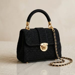 Könnte beinhalten: Schwarze Häkeltasche mit goldfarbener Kette und Beschlägen. Die Tasche hat einen oberen Griff und einen Klappenverschluss mit goldenem Verschluss. Die Tasche ist ein strukturiertes Design, geeignet für verschiedene Anlässe.