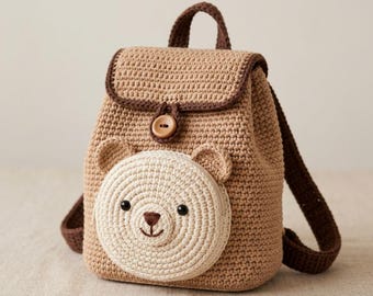 Puede incluir: Una mochila de ganchillo color canela con un bolsillo frontal circular en forma de cara de oso color crema. La bolsa tiene bordes marrón oscuro, un asa superior, dos correas para los hombros y un cierre de botón de madera en la solapa.