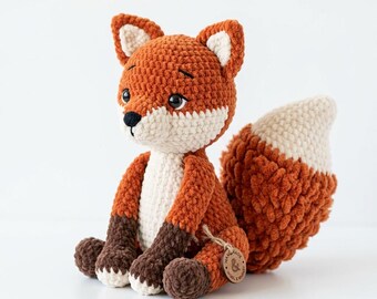 Puede incluir: Un peluche de zorro tejido a ganchillo con un cuerpo de color naranja vibrante, pecho y cara de color crema, y patas de color marrón oscuro. El zorro tiene ojos negros grandes, una nariz negra pequeña y una cola grande y tupida con la punta crema. Tiene una etiqueta de madera.
