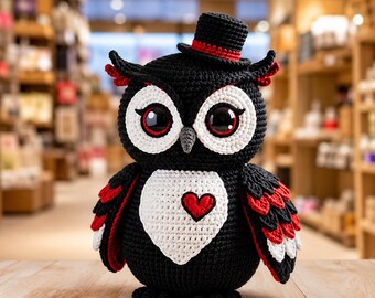 Puede incluir: Una figura de búho tejida a ganchillo con hilo negro y detalles blancos alrededor de los grandes ojos y en el pecho. El búho lleva un pequeño sombrero de copa negro con una banda roja y tiene un corazón rojo cosido en su pecho blanco. Las alas muestran plumas superpuestas negras, rojas y blancas.