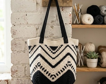 Puede incluir: Un bolso tote de ganchillo con un patrón de chevrón en negro y crema y dos asas negras. El bolso está confeccionado con un punto de hilo texturizado y muestra un diseño geométrico que transiciona desde una parte superior crema a una base negra.