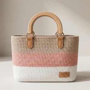 Puede incluir: Bolso tote de ganchillo hecho a mano con asa de madera y detalles de cuero. El bolso presenta un diseño de bloques de color con rayas beige, rosa y blancas. Una pequeña etiqueta rectangular con el texto "HANDMADE BLOSSOM & THREAD" está adjunta.