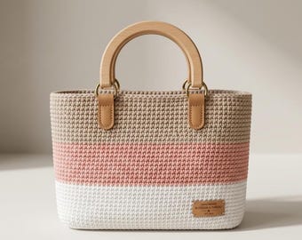 May include: A handmade crochet tote bag with a wooden handle and leather accents. The bag features a color block design with beige, pink, and white stripes. A small rectangular tag with the text "HANDMADE BLOSSOM & THREAD" is attached to the front.