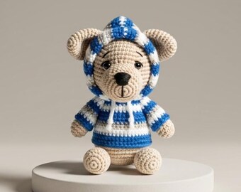 May include: A crocheted teddy bear wearing a blue and white striped hoodie and matching sweater. The bear has tan fur, a black nose, and is sitting on a white circular platform. The handmade toy is a charming example of crochet artistry.