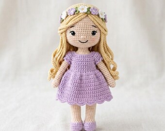 Puede incluir: Una muñeca de ganchillo hecha a mano con largo cabello de lana rubia, que lleva un vestido color lavanda con dobladillo festoneado y una corona de flores blancas y moradas. La muñeca tiene tez beige claro, ojos bordados negros y una sonrisa.