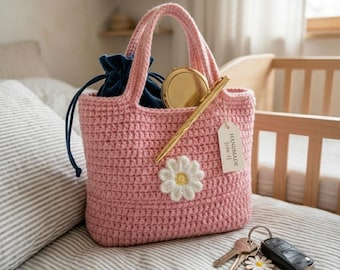 May include: A pink crocheted tote bag featuring a white daisy appliqué on the front. The bag contains a dark blue velvet drawstring pouch, a gold compact mirror, and a gold pen. A white tag labeled HANDMADE [cite: 1] hangs from the handle.