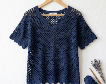 May include: A dark navy blue short-sleeved crochet top featuring an openwork pattern, a notched neckline, and scalloped edges. A label inside the collar reads "HANDMADE BY SEA & STITCH". A ball of matching yarn and a wooden crochet hook are nearby.
