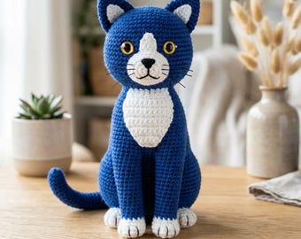 Puede incluir: Una figura de gato tejida a ganchillo con hilo azul oscuro para el cuerpo y blanco para el pecho, las patas y el interior de las orejas. El gato tiene grandes ojos amarillos, nariz y boca bordadas en negro y bigotes negros sobre el hocico.