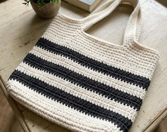 May include: A crochet tote bag featuring a repeating pattern of horizontal cream and charcoal gray stripes. The bag is constructed with a textured yarn stitch and includes two integrated handles at the top, maintaining a consistent striped design throughout the body.