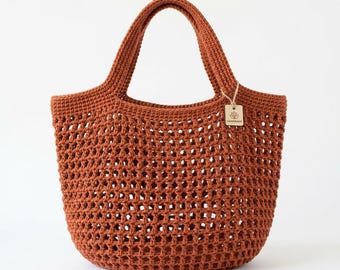 Puede incluir: Un bolso de ganchillo color óxido con un patrón de malla abierta en todo el cuerpo y dos asas resistentes. Una pequeña etiqueta rectangular de color beige con la palabra "HANDMADE" y un icono de flor está sujeta al asa derecha con un cordel.