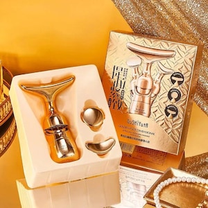 Puede incluir: Un juego de masajeador facial dorado, que incluye una herramienta principal y dos accesorios más pequeños, presentado en una bandeja de color crema. El embalaje presenta detalles y texto dorados en relieve, con las palabras "Golden Four Dimensional".