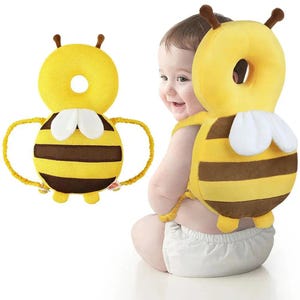 May include: A yellow and brown bee-shaped baby head protector. The head protector has a circular design with a hole in the center and two brown antennae. The body of the bee has brown stripes and white wings. The protector is worn by a baby.