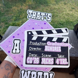 Può includere: Un cappello di laurea con un design glitterato viola, con le parole "THAT'S" e "WRAP!" in bianco. Un design di ciak include il testo "Production: Graduation" e "Director: AUNDREA."