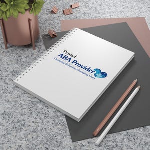 May include: A white spiral-bound notebook with the text "Proud ABA Provider" in blue. The notebook also features a blue and white graphic and the text "Changing Behavior, Changing Lives". Two pencils rest beside the notebook.